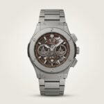 Silver-tone chronograph watch with brown skeleton dial, three sub-dials, tachymeter bezel, and stainless steel bracelet.