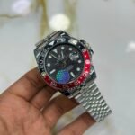 Silver-tone GMT style watch with black dial, red and black bezel, luminous markers, and jubilee bracelet held in hand.
