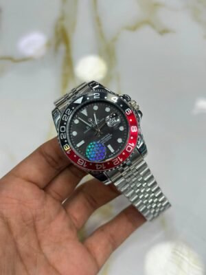 Silver-tone GMT style watch with black dial, red and black bezel, luminous markers, and jubilee bracelet held in hand.