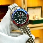 Silver-tone GMT style watch with black dial, red and blue rotating bezel, and jubilee bracelet held in hand.